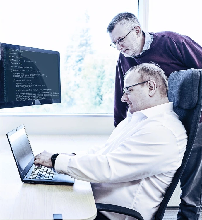 Two men working together at a desk with a laptop and monitor displaying code, in a modern office setting with a window and shelving unit nearby. One man sits in a chair while the other stands behind him, collaborating on a project.