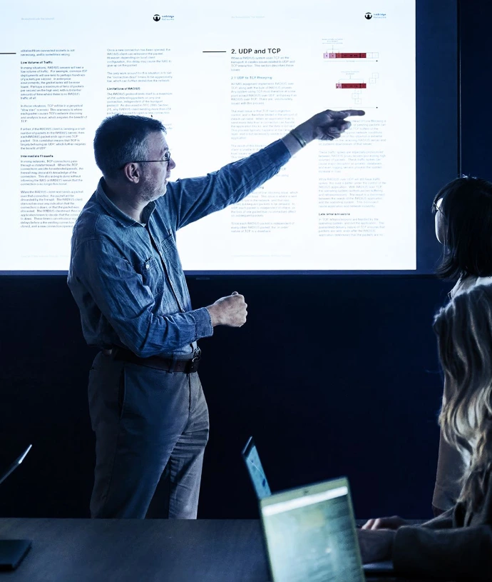 Man presenting to audience in meeting room, pointing at projected screen with text and charts, with people seated at table with laptops, engaged in professional discussion or training session.