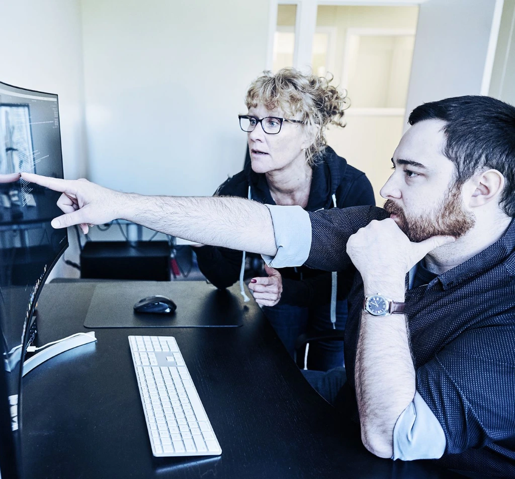 Two men in office setting, one pointing at computer screen, discussing data, wearing glasses and watch, sitting at desk with keyboard and mouse.