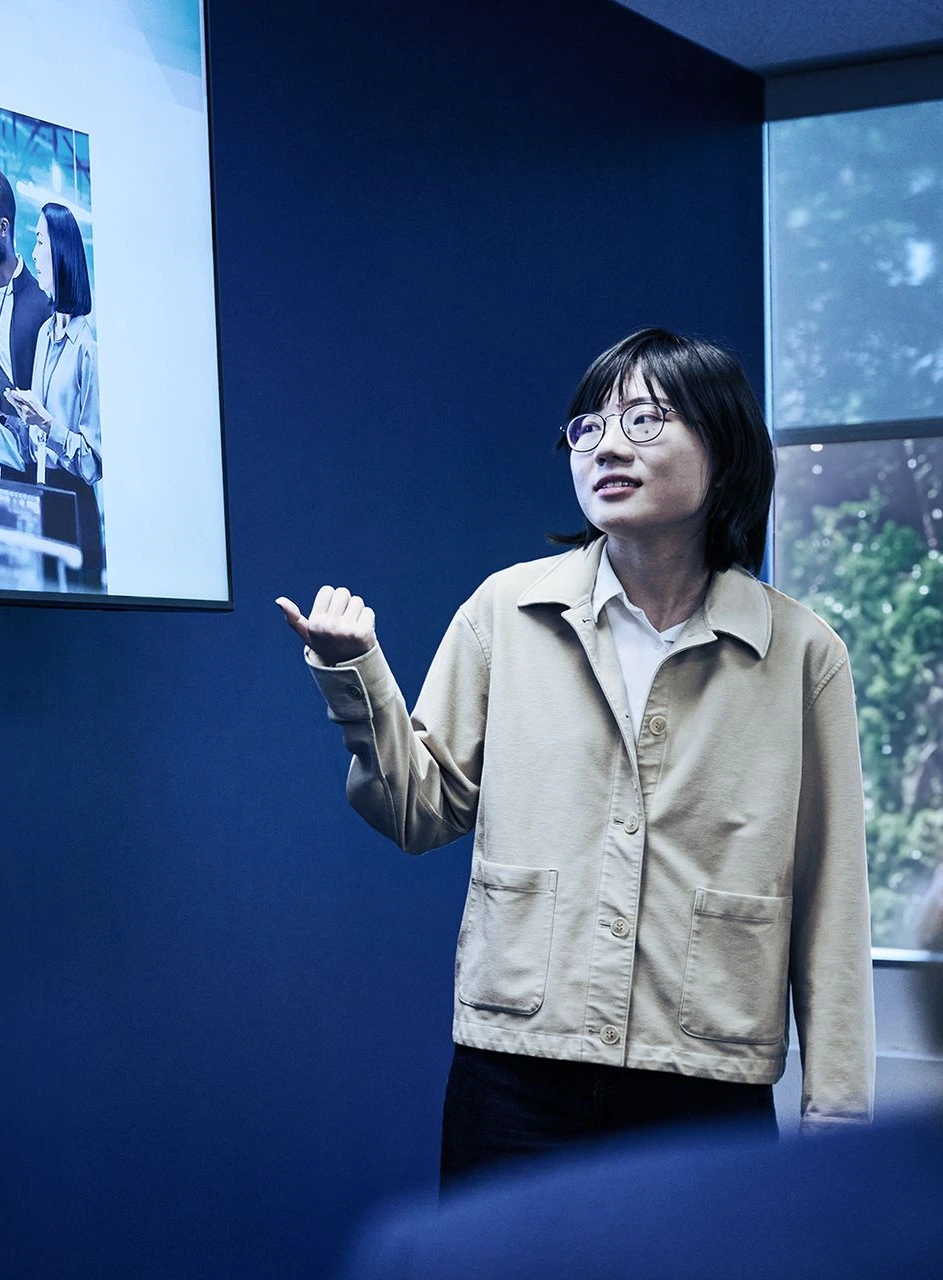 Woman presenting to a man in front of a screen with presentation content and photo of people interacting, standing in blue room with window showing trees outside, discussing device authentication and TLS session.