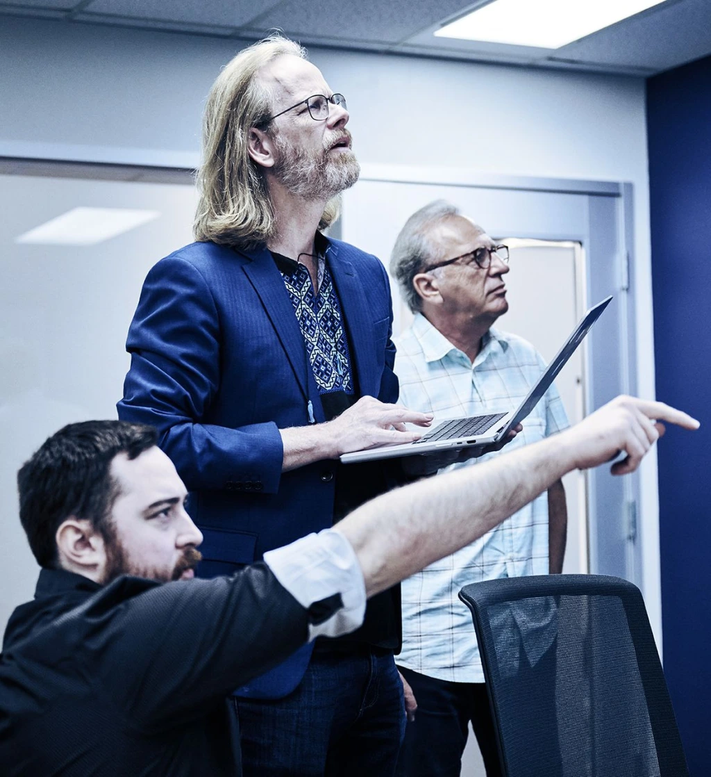 Three men in an office setting, one holding a laptop and pointing, another seated and pointing, and a third standing beside them, all looking up and engaged in discussion.
