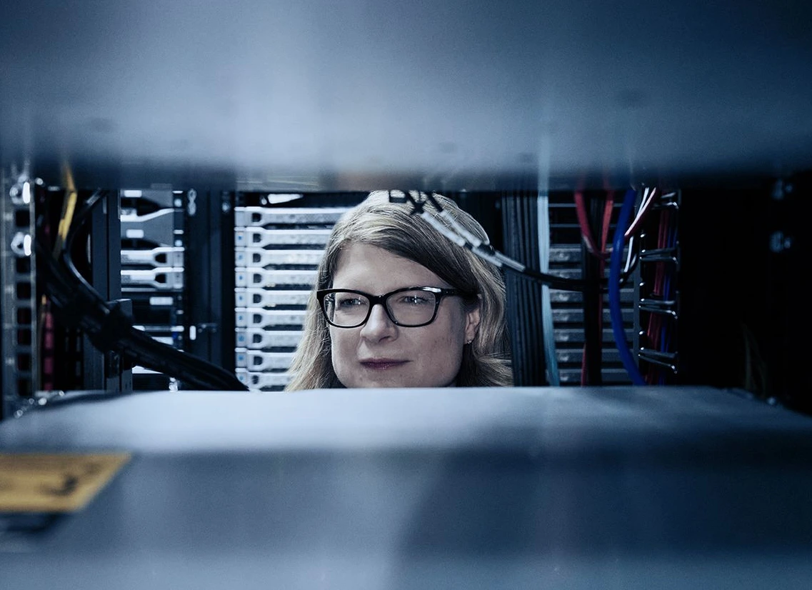 Woman with glasses working on computer in server room, surrounded by rows of computer servers and cables, focused on screen.