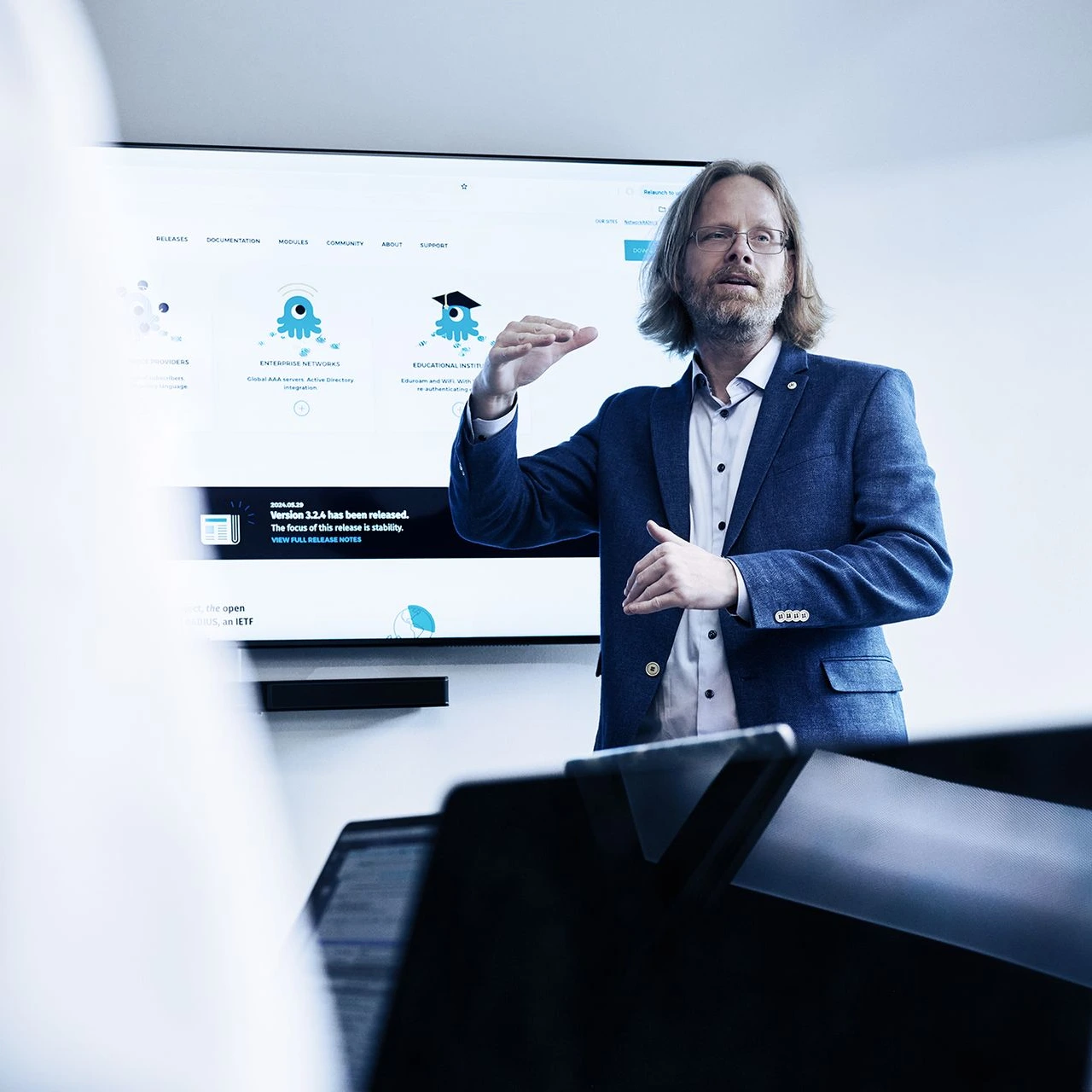 Businessman in blue suit presenting to audience in front of large screen display with educational icons and text, gesturing with hands in professional setting.