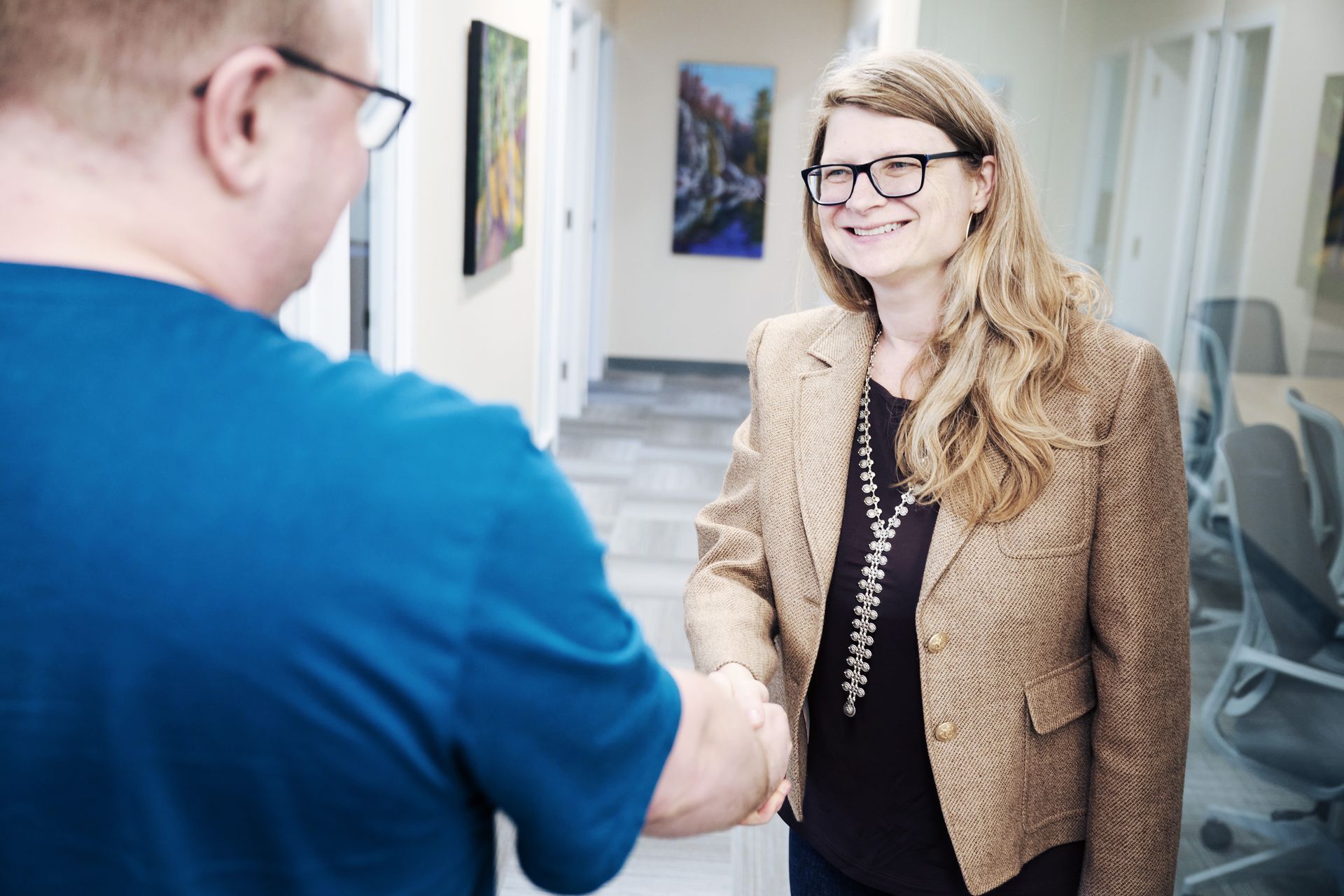Businesswoman and man shaking hands in office hallway, exchanging greetings. Woman wears brown blazer and black shirt, man wears blue shirt. Office setting with paintings on wall and chairs in background. Professional networking and meeting concept.