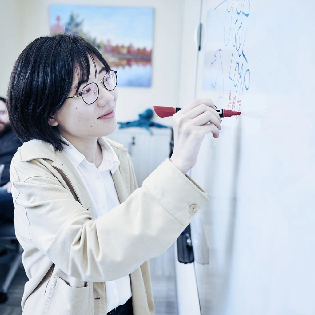 Woman in beige coat writing on whiteboard with red marker, standing in office meeting room with colleagues seated at table in background, surrounded by modern decor including painting on wall.